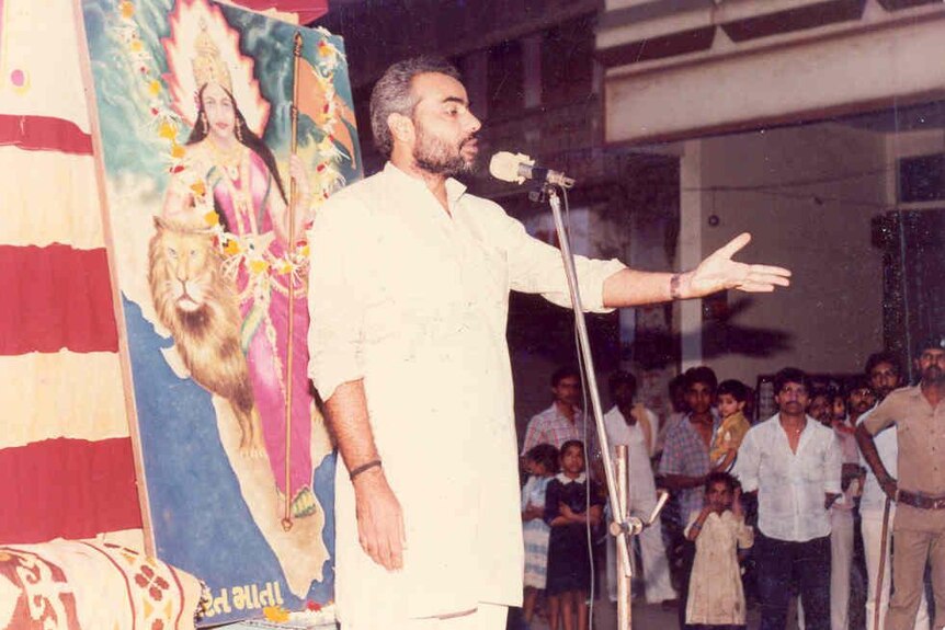 Narendra Modi as a young man stands at a microphone, extending a hand out while speaking to a crowd
