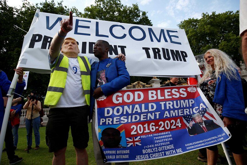 A man wearing a Donald Trump mask stands in front of a sign saying "Welcome President Trump".