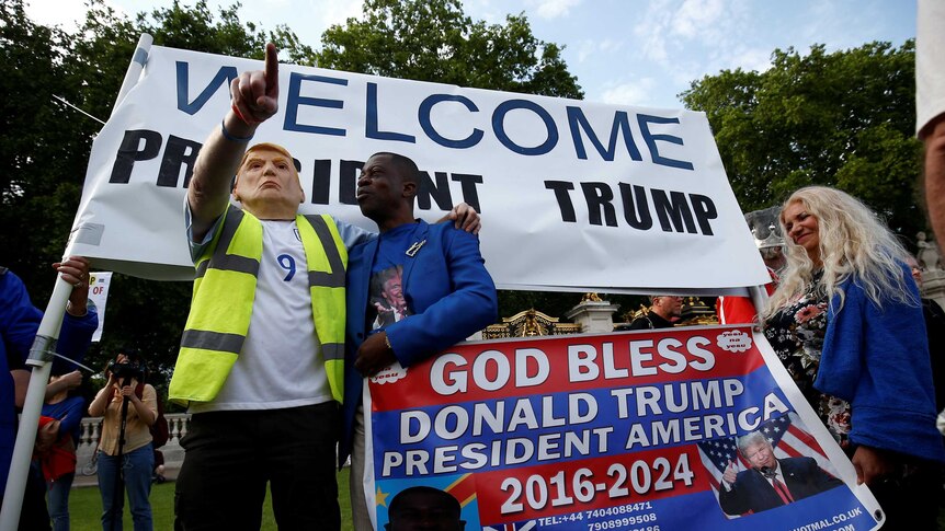 A man wearing a Donald Trump mask stands in front of a sign saying "Welcome President Trump".