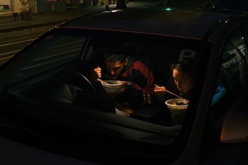 A young man and woman sit in the front seats of a car eating takeaway.