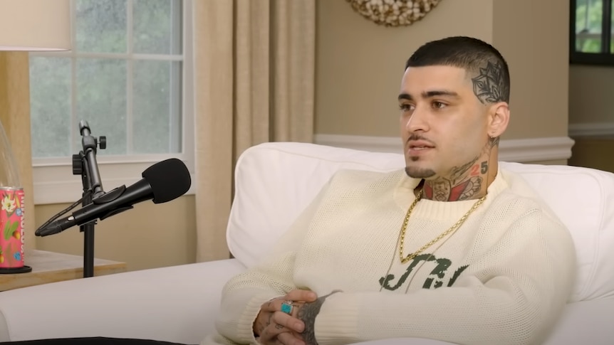 A man wearing a cream-coloured sweater sitting on a white sofa inside a house