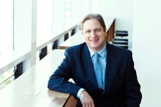 A man in a blue suit and tie sits at a desk, smiling at the camera.