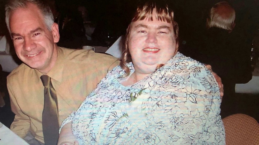 Debbie and her husband Ian sit at a table dressed up and smiling at the camera, Ian has his arm around Debbie.