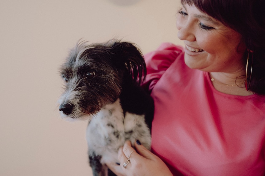 A woman with dark hair and a pink top is smiling down at the small dark haired dog she is holding. 