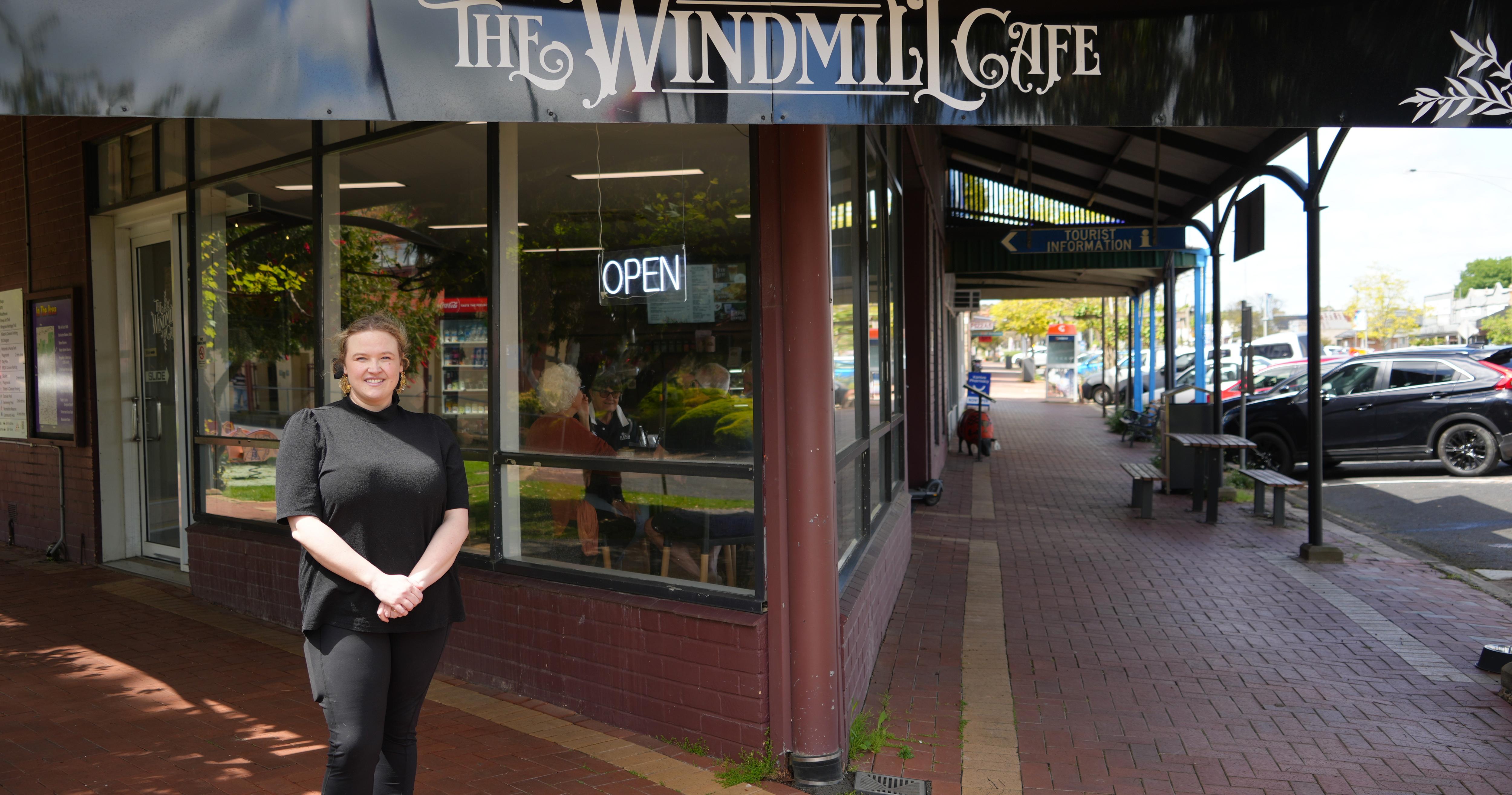 A woman stranding in front of a corner building, bearing the sign "The Windmill Cafe" on the large window.
