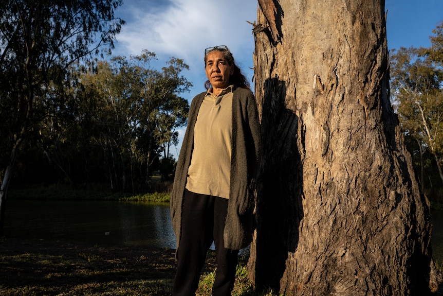 A woman stands against a gumtree along a riverbank.