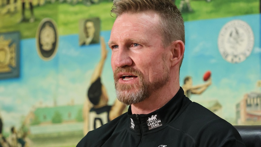 Nathan Buckley, wearing a Collingwood-branded jacket, speaks.  