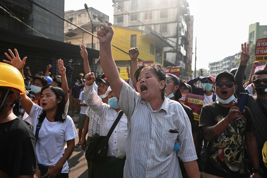 A middle-aged woman with short hair and striped shirt raises her fist and chants in crowd in city.