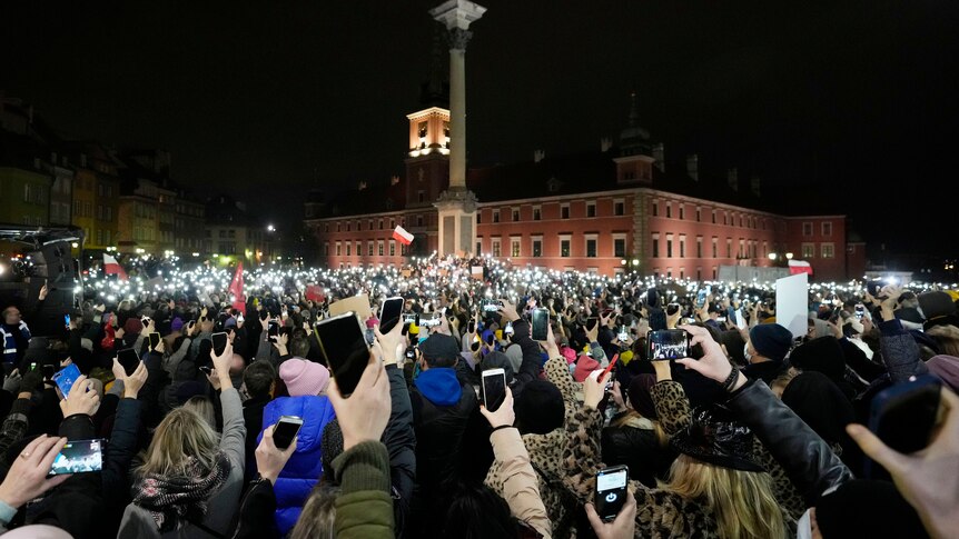 A large group of calm protesters face an offical building and light up their mobile phone torches and hold their devices aloft.