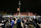 A large group of calm protesters face an offical building and light up their mobile phone torches and hold their devices aloft.