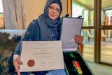 A woman in a headscarf holds up a medal and a university degree