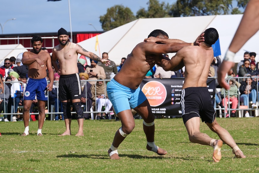 Two men without tops wrestle on a sports field with onlookers and a crowd