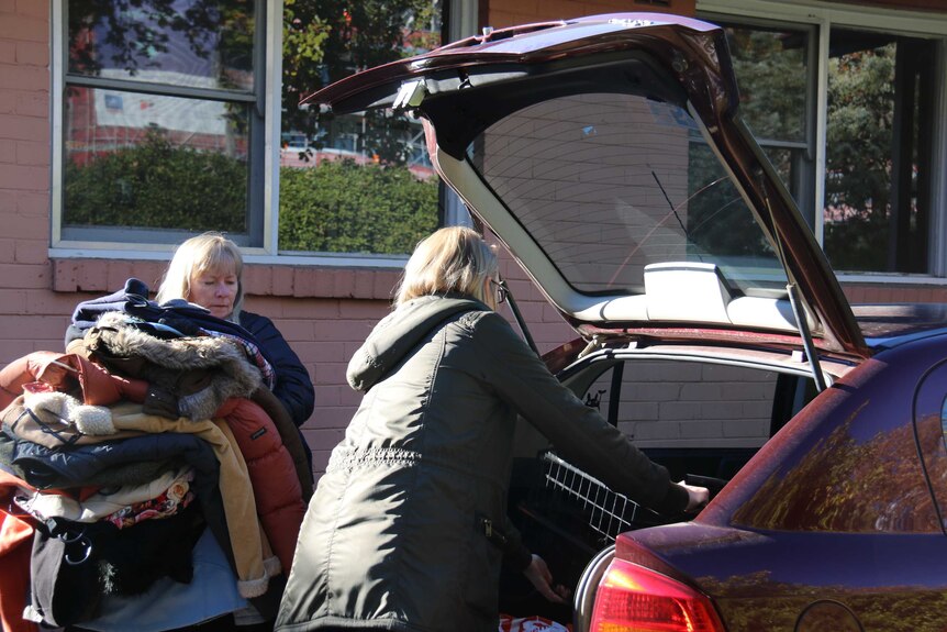 Two women pick up dozens of coats and put them into the boot of a car.