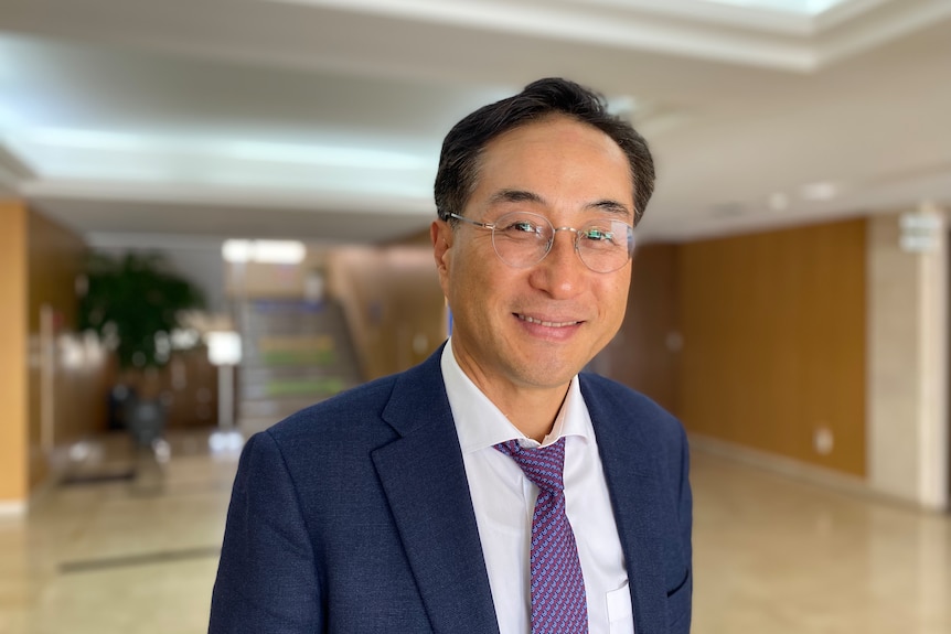 A middle-aged Korean man wearing a suit and tie and glasses smiles in a building foyer.