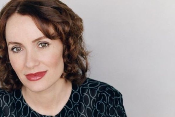 A close shot of a woman with brown hair, dark print top and red lipstick, smiling slightly with closed mouth.