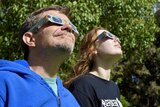 A man and a woman wearing eclipse glasses look in the direction of the sun.