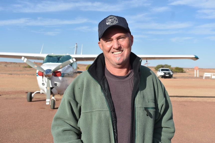 An man wearing a hat and green jumper stands in front of an airplane.