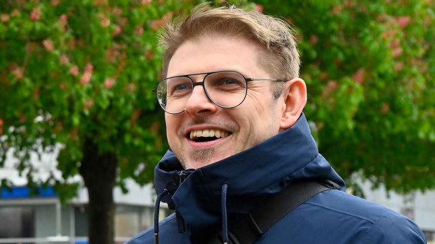 A smiling middle-aged man with glasses and a blue jacket stands outside.