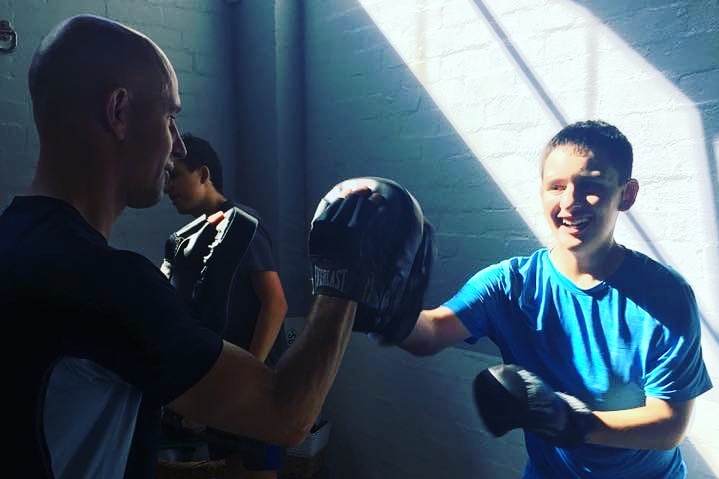 Oliver Moore wears boxing gloves and swings at boxing pads held by an older man