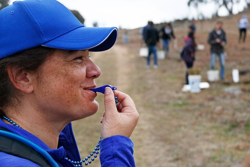 Lady wearing a blue cap blows on a blow whistle. People can be seen planting in the background.