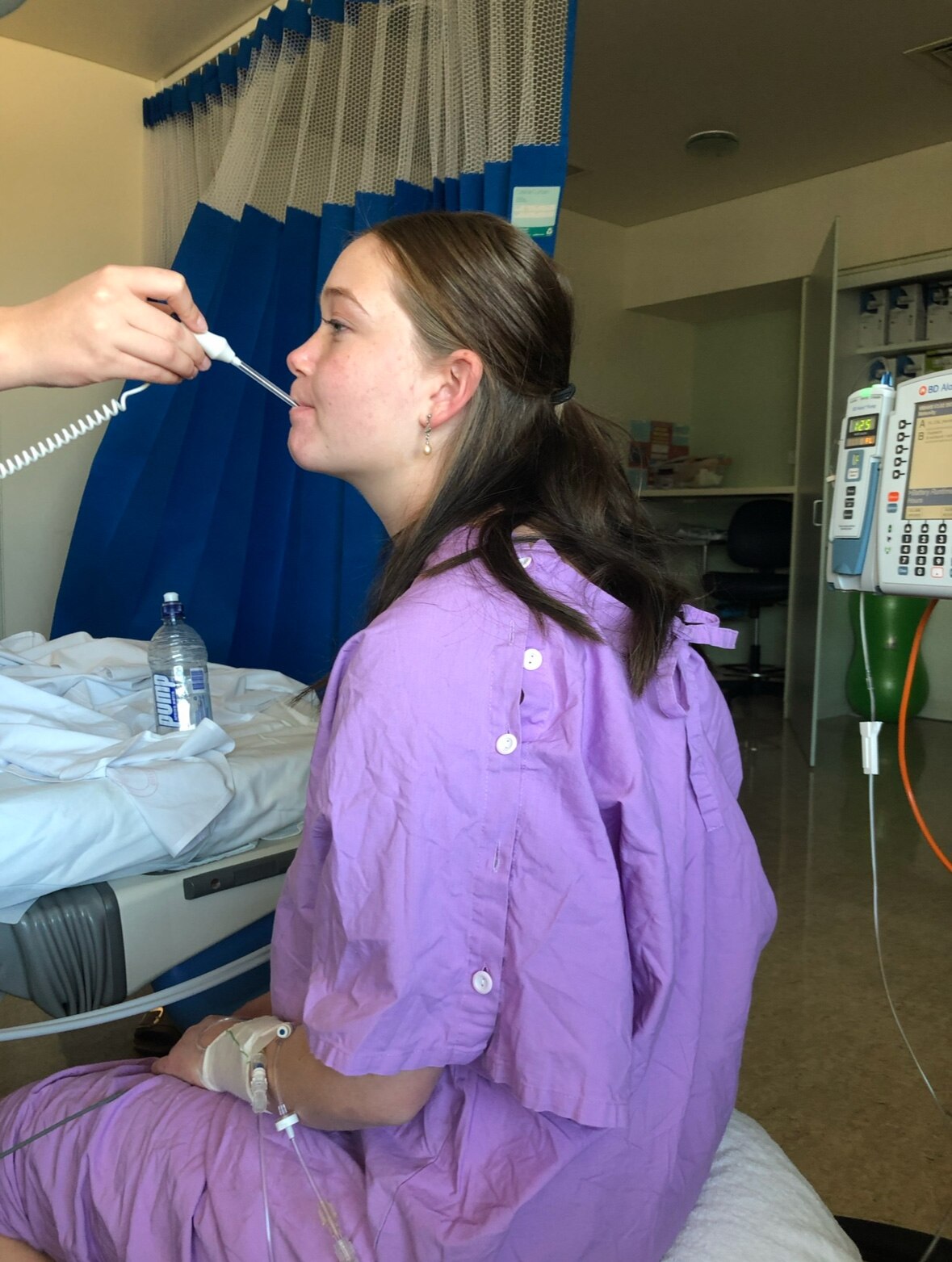 A young woman wearing a purple hospital gown sits on a bed with a thermometer in her mouth, the hospital room in the background