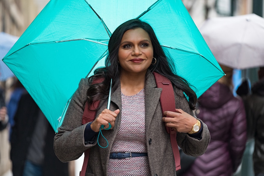 A woman wearing corporate attire stands on a city street and smiles while holding an open turquoise folding umbrella.