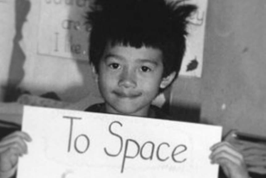 A six year old boy holding a poster in a classroom that reads "To Space."