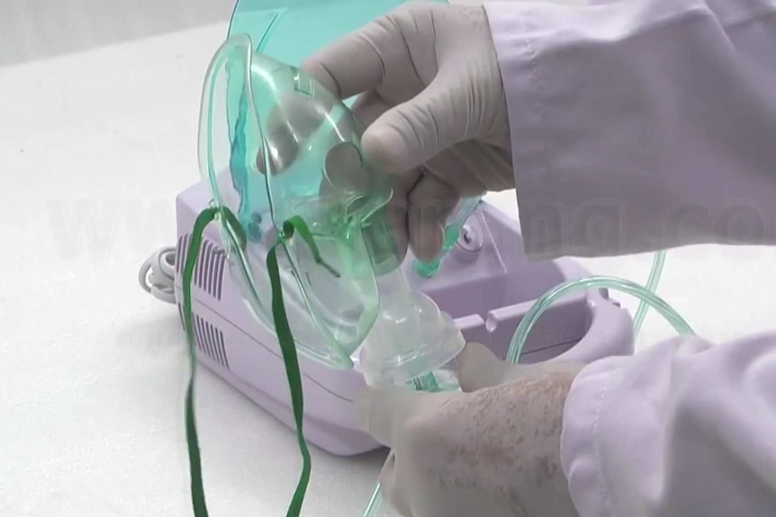 Two hands on a plastic facemask with tubes attacked with a white device in the background.