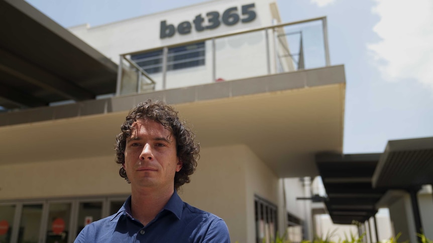 A man in a blue polo shirt stands in front of a beige office building, with the word bet365 on it.