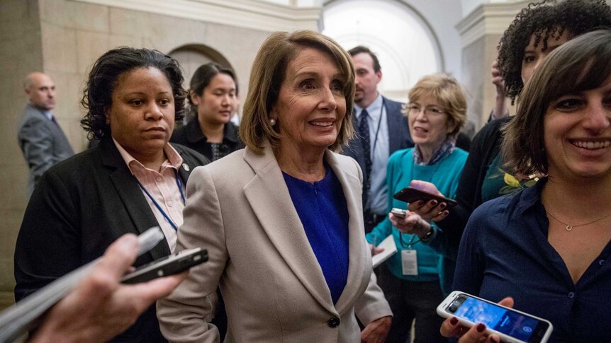 Nancy Pelosi wears a tan coat and blue top and is seen smiling and surrounded by eight reporters near Capitol building.