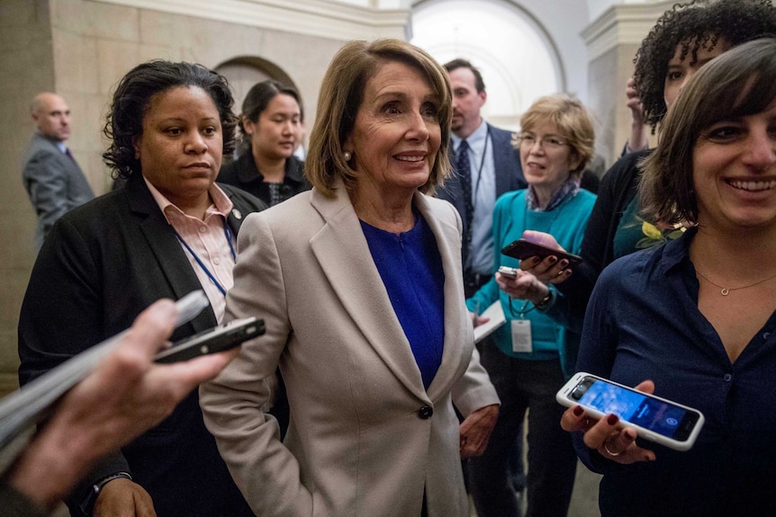 Nancy Pelosi wears a tan coat and blue top and is seen smiling and surrounded by eight reporters near Capitol building.