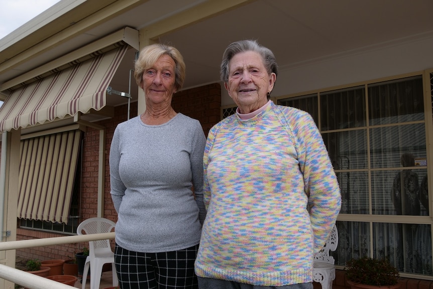 Two older women stand side by side out the front of a house./