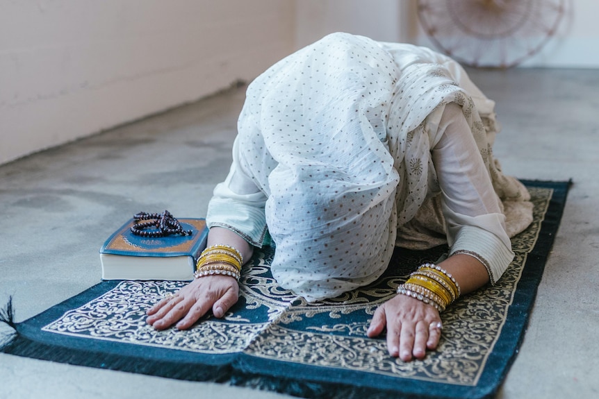 Muslim woman wearing headscarf in prayer position on prayer mat.