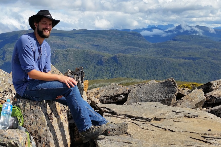 A man wearing a long sleeved blue shirt, blue jeans and a hat sits on a rock overlooking a national park.