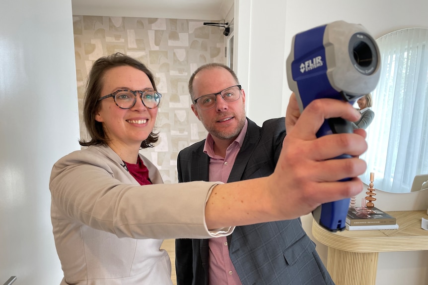 A man and woman standing in a home bathroom hold out a thermal camera.