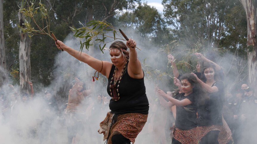 Female dancers wave branches as they dance through smoke in front of a crowd at an event in a nature reserve.