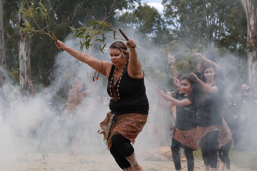 Female dancers wave branches as they dance through smoke in front of a crowd at an event in a nature reserve.