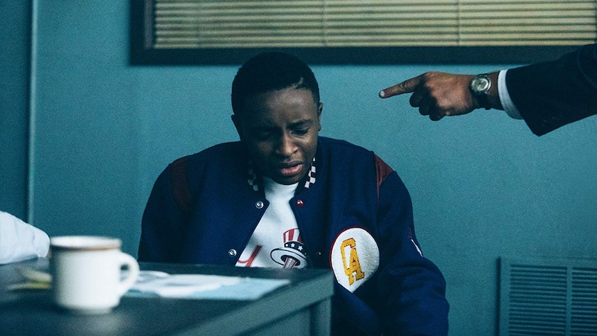 A police officer points at a young, black man in an interrogation room.