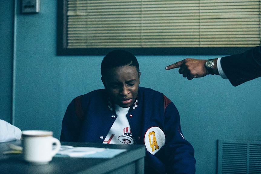 A police officer points at a young, black man in an interrogation room.