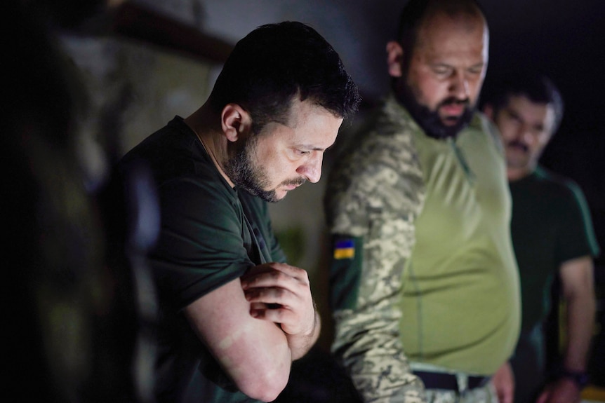 Volodymyr Zelenskyy (left) leans over a railing while standing next to a man in dressed in army fatigues.