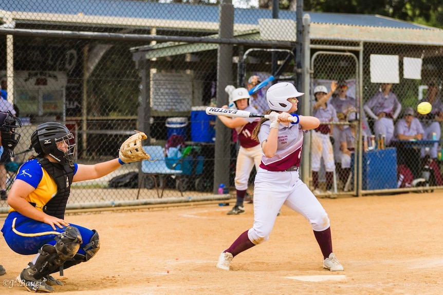 A softball batter wearing a white helmet and maroon and white uniform waits for the ball with her bat with a catcher behind.