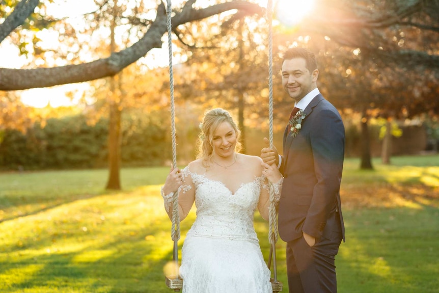 A young woman in a wedding dress sits on a swing bathed in golden light while her husband smiles happily at the camera.