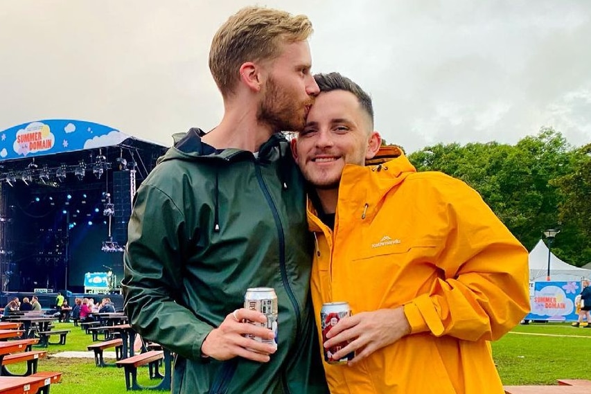 James kisses Tom on the head as they stand at a sodden outdoor concert, wearing raincoats.