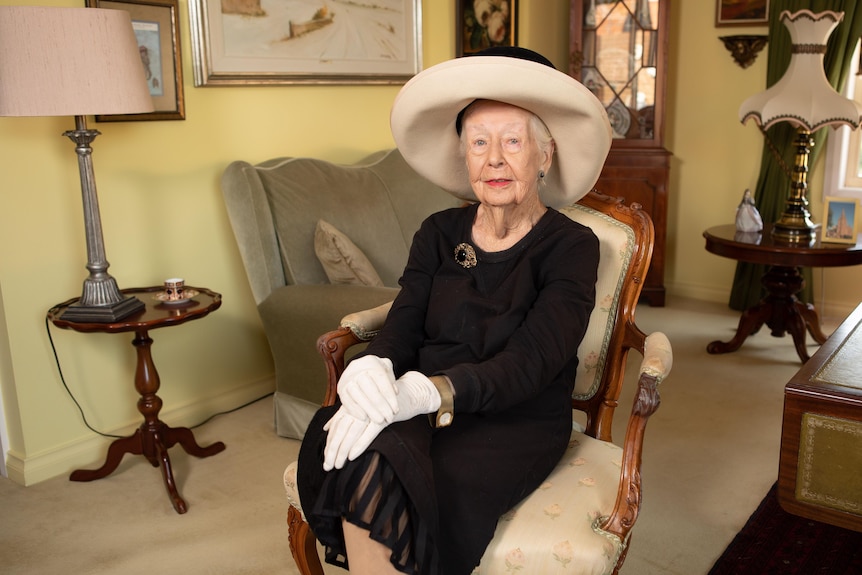 A woman in fine traditional clothing, including a wide-brim hat and gloves, poses on a chair for a photo.