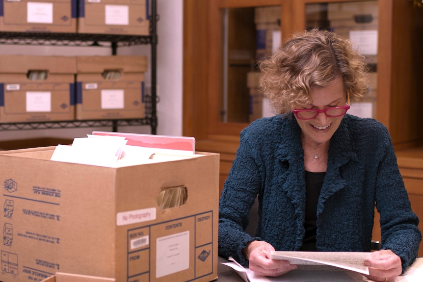 The author Judy Blume - a woman in her mid 80s with curly red hair and blue glasses - sits and reads a letter in an archive room