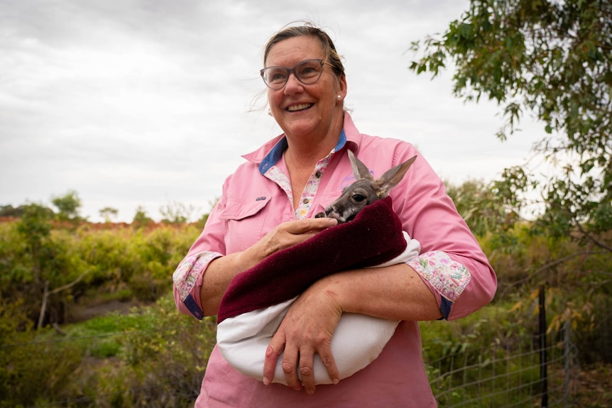 A woman in a pink shirt stands and smiles while she cradles a joey kangaroo in a cloth pouch in her arms.