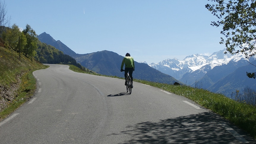 A photograph of a cyclist riding through the mountains in France, with snow capped mountains in the background