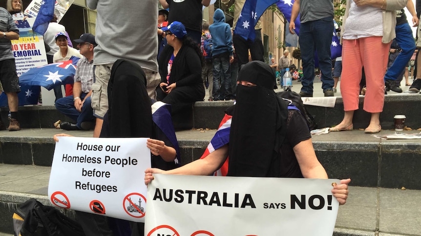 Two people wear niqabs while holding anti-refugee and anti-Islamic signs.