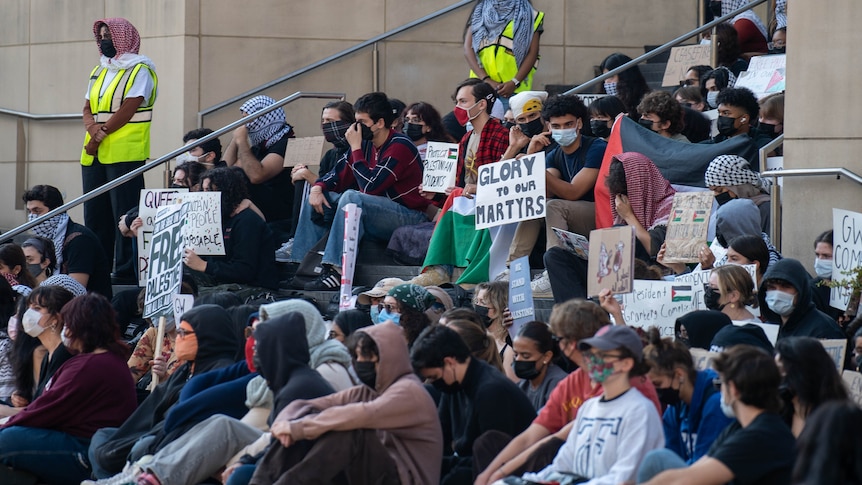 A group of students sits on outdoor stairs. Some hold signs with messages like 'glory to our martyrs'.