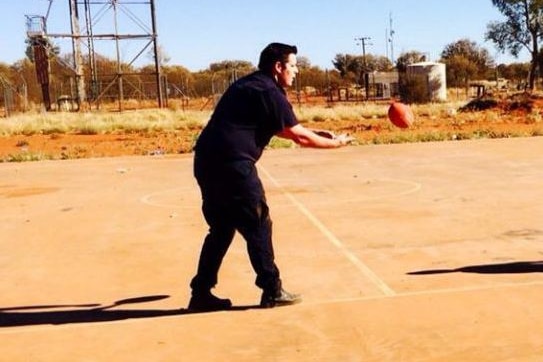 A man in uniform plays footy on a dusty basketball court